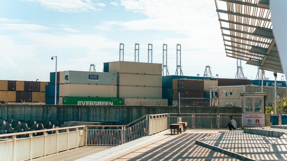 View of a shipping port with stacked containers and a man resting nearby under industrial shadows.