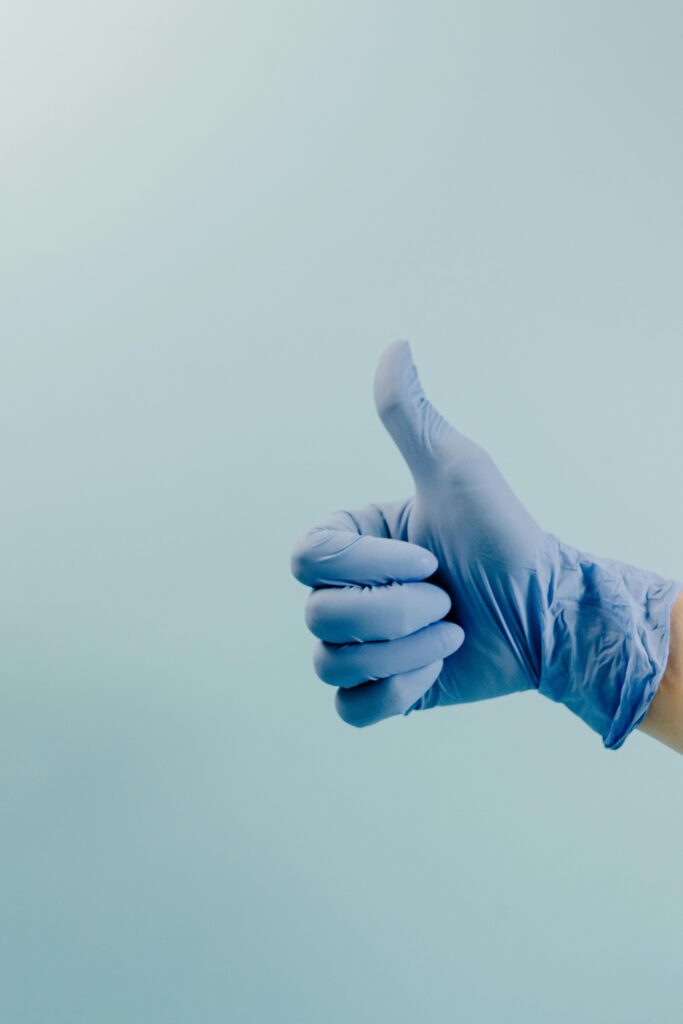 A blue surgical gloved hand showing a thumbs up gesture against a cyan background.